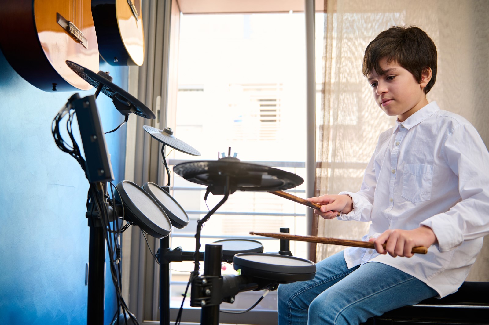 Caucasian teenage boy beating with drumsticks on the cymbals while performing sound on the drum kit, playing music in his room with guitars hanging on the blue wall